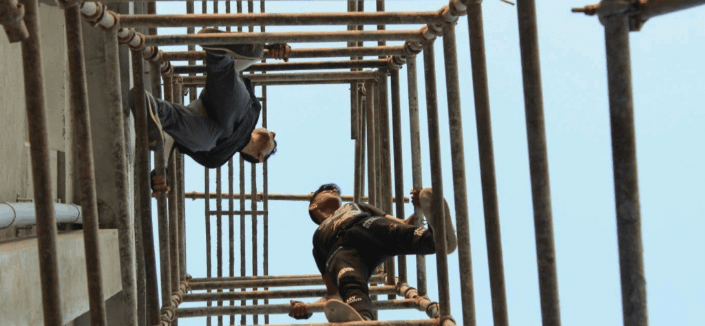 Looking up at two men standing on some bars high in the air (unsure how to describe this one accurately!)