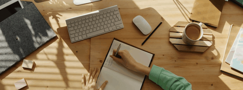 overhead photo of someone at a desk writing in a notebook - their computer is not far away.