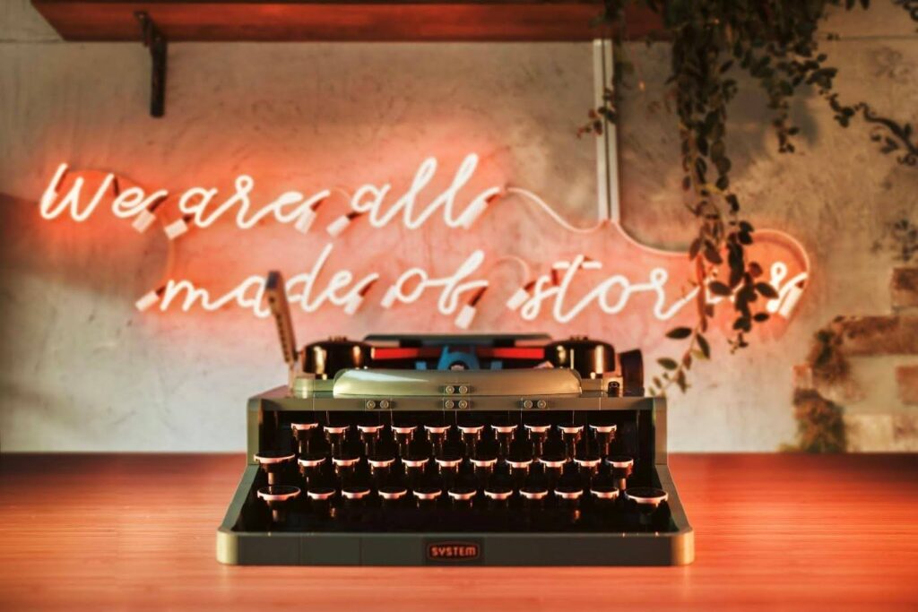 A typewriter sits in front of a neon sign that reads "we are all made of stories"
