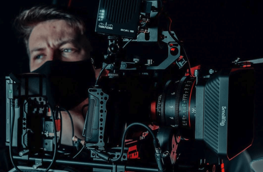 A photo of a man wearing a mask (medical) and holding a camera, presumably on the set of a film or a tv show.