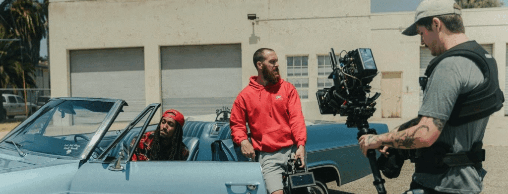 Two guys in a convertible being filmed by a third guy. It's either a music video or perhaps a fashion commercial.