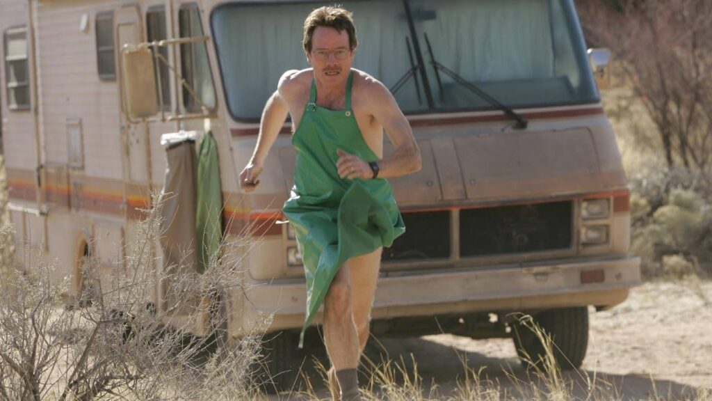 Walter White in nothing but boots and a green apron runs away from a crappy old RV