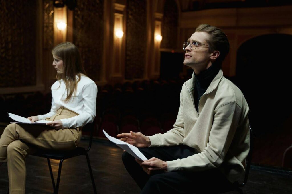 Two actors rehearse a play on stage, each holding a script.