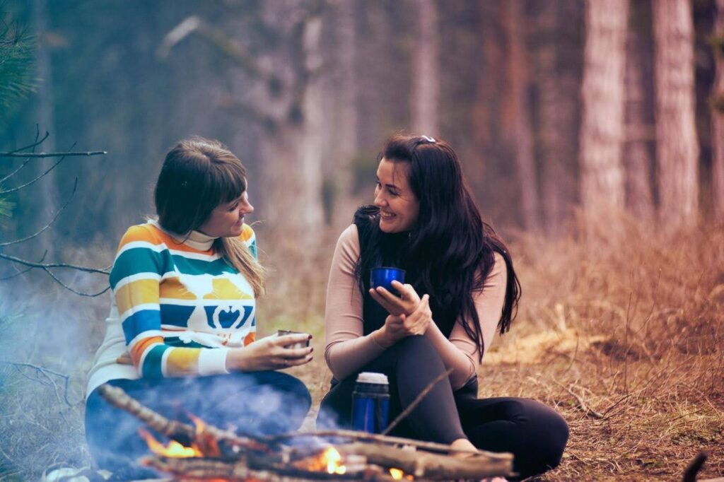 Two women camping in the forest, sitting in front of a fire, drinking a hot beverage.