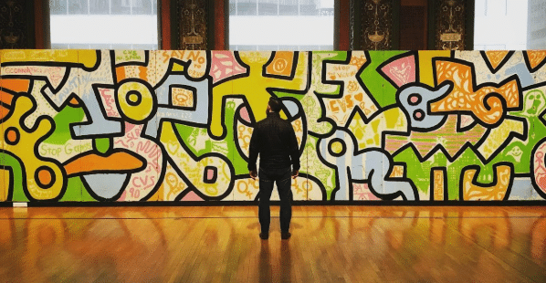 A photo of a man standing in front of a wall of colorful artwork, taking it all in.