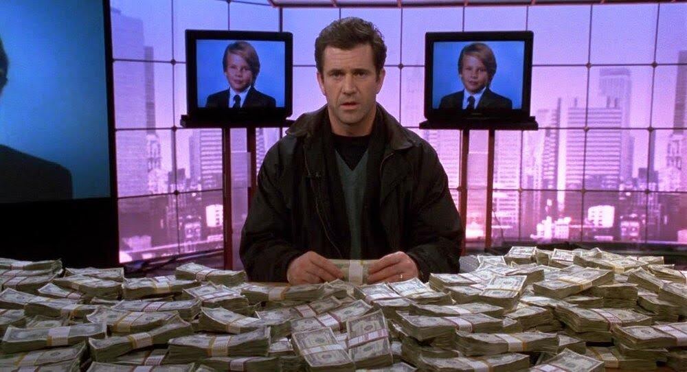 A still from the 1996 movie Ransom. In the photo you see Mel Gibson's character sitting at a table covered in money with photos of his missing son on two televisions in the background.