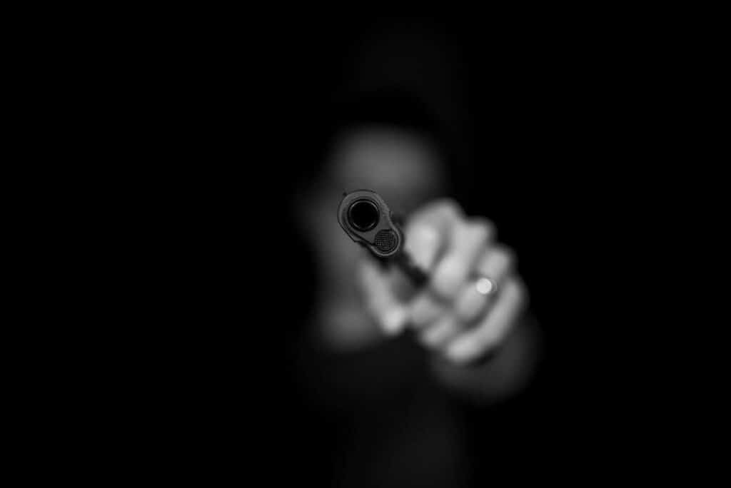 This is a photo implying a POV shot in film. You see the barrel of a gun in focus, and a hand holding it out of focus.