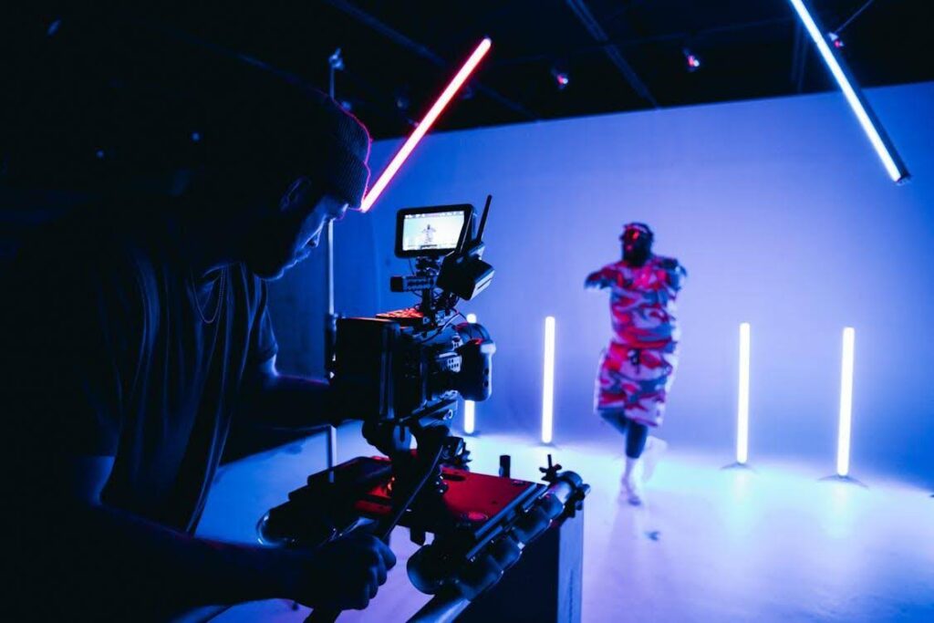 A cinematographer on set, setting up lighting, which is one of their responsibilities. A man in red and black camo stands under the lighting, being filmed.