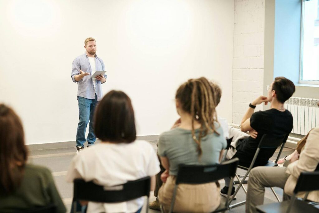 A man speaks in front of a room of people who are seated in folding chairs.