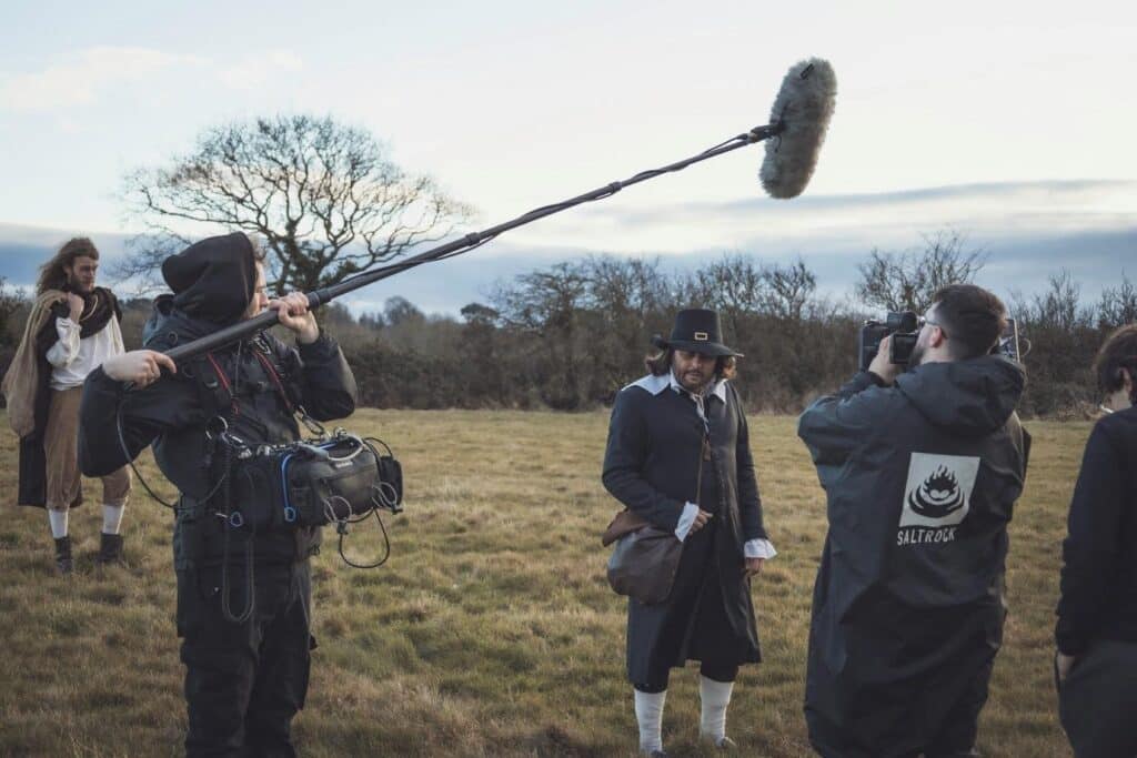 A film crew films an outdoor scene - a period piece (men are dressed as what appear to be pilgrims) - a boom audio arm hangs overhead
