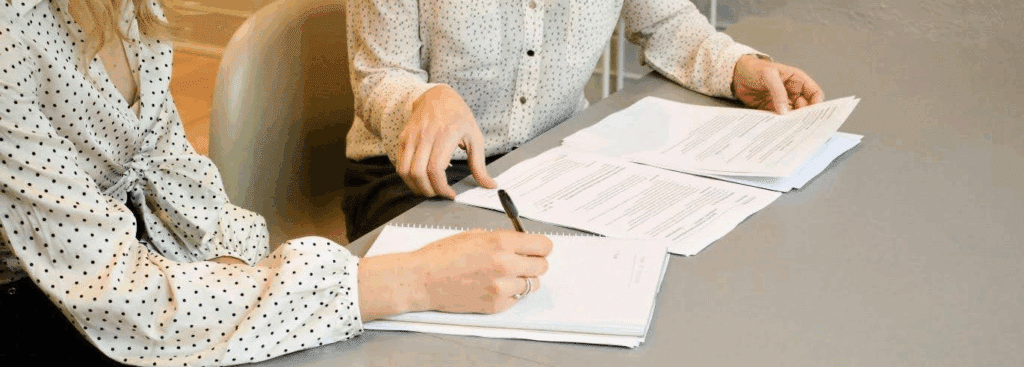 Two people sit at a table, one is looking over a contract and the other is taking notes.