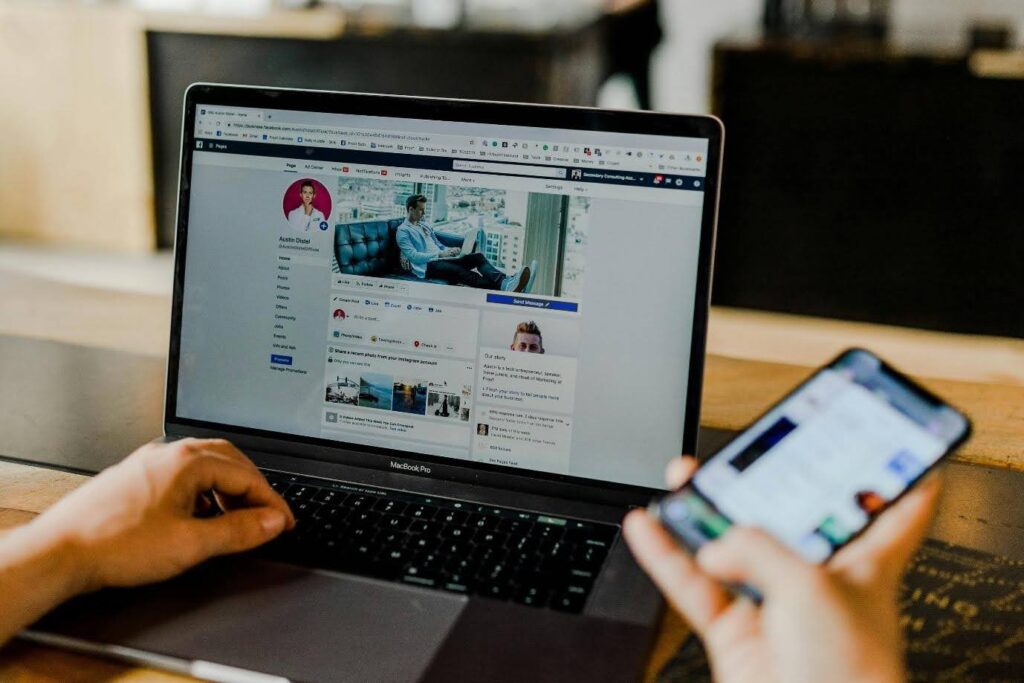 Photo of someone sitting at a laptop with a Facebook profile on their screen and a smart phone in hand.