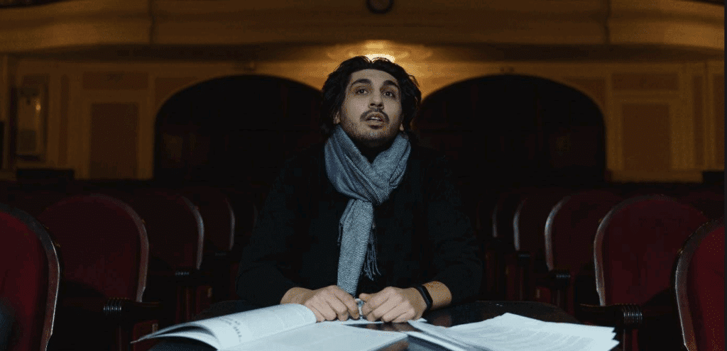 A playwright sits in the center of a theater, looking toward the stage, surrounded by play scripts