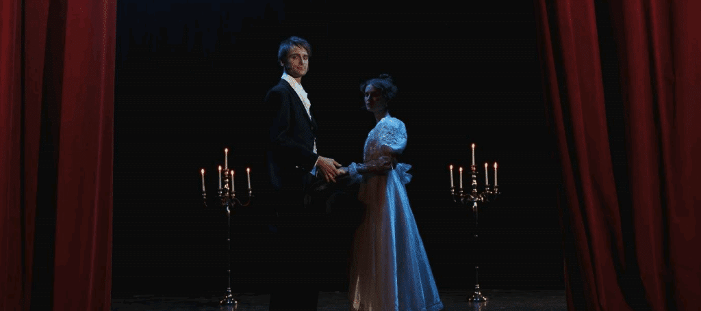 Two actors in victorian costumes standing in the middle of a stage, framed by red curtains