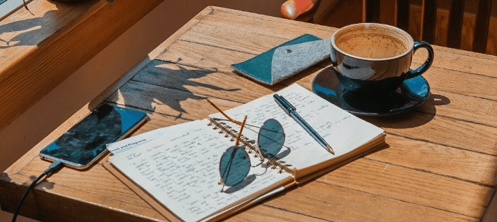 a small table in a window. on top sits a cup of coffee, an open notebook with a pen, sunglasses, and a cell phone charging.