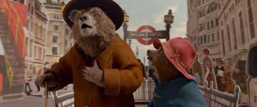 Paddington, wearing a red hat, talks to another bear in front of Piccadilly Circus.