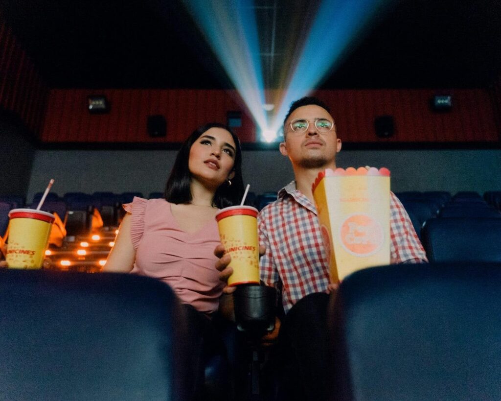 A woman and a man sit side by side in a movie theater, cups and popcorn in hand.