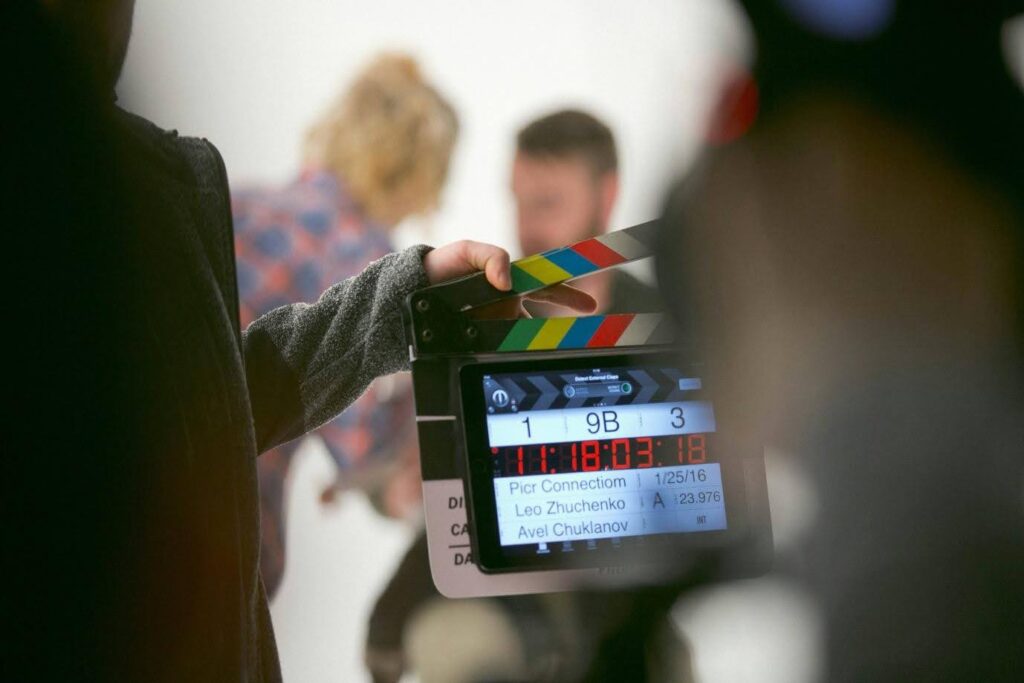 In focus is a film production clapboard - unfocused in the background is at least one actor (there are two people, but the 2nd might be a makeup/wardrobe/hair person)