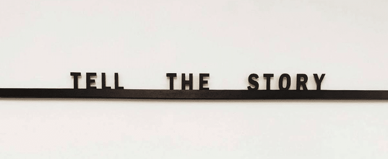 Sort of looks like a black shelf on a white wall with wooden letter blocks that have been put together to read "TELL THE STORY"