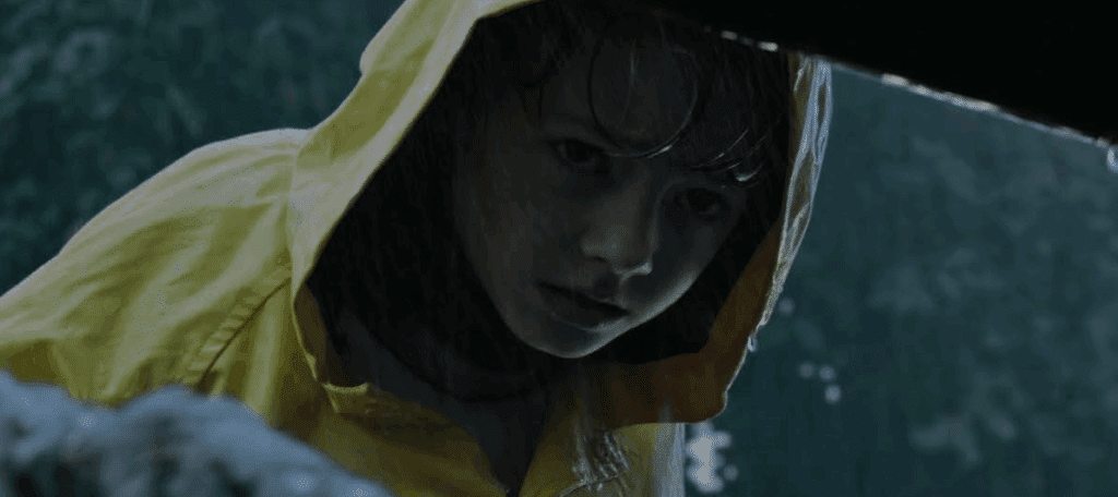 Georgie, wearing a yellow raincoat, peers down the sewer drain