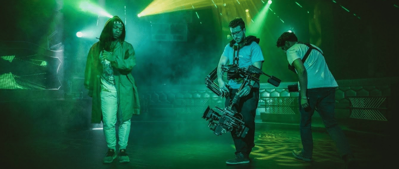 Part of a film crew is visible (a gaffer and a cameraman) film what looks like a music video, surrounded by green lighting.