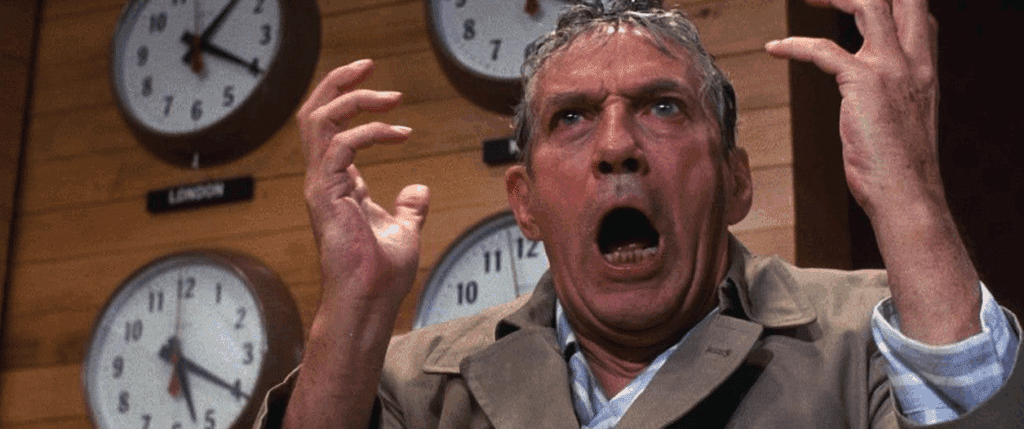 an older reporter (man) looking pretty crazy with a bunch of clocks hanging behind him on a wood-paneled wall in 1979's Network